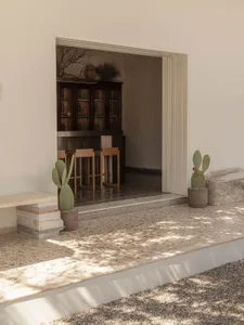 View from a sunlit terrazzo outdoor terrace with potted cacti and stacked stone bench, looking through an open doorway into a dark wood bar area with wooden stools.
