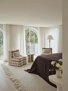Bright modern bedroom featuring arched windows with sheer curtains, two striped linen armchairs, a large bed with chocolate brown linen coverlet, and a cream shaggy rug on light wood flooring.