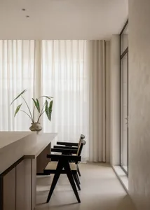 Modern minimalist dining area featuring a light oak counter, three black wooden dining chairs with woven seats, a potted plant, and floor-to-ceiling sheer white curtains.