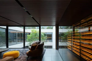 Modern living room featuring a dark wood slatted ceiling, large glass walls overlooking a garden, brown curved sofa, bright yellow ottoman, and illuminated wooden shelving unit.
