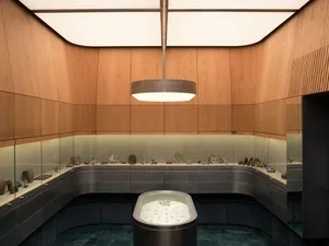 Modern luxury jewelry store interior featuring curved walnut wood paneling, dark metal display cases, glass vitrines, and a central oval island showcase under a large circular pendant light.