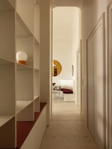 View down a minimalist cream-colored hallway featuring built-in shelving with red accents, leading to a curved dining nook with a red upholstered bench and transparent chairs.