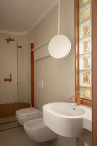Modern minimalist bathroom with terracotta accents, white wall-hung toilet and bidet, integrated white ceramic sink with matching orange faucet, round mirror, and glass block window.