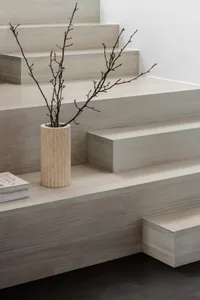 Close-up of a minimalist wooden staircase with light oak treads and risers, featuring a cylindrical travertine vase holding bare branches on a step, next to a stack of books.