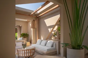 Bright Mediterranean-style courtyard lounge featuring a built-in concrete sofa with white cushions, wooden hanging lanterns, large potted plants, and dramatic sunlight patterns on beige walls.
