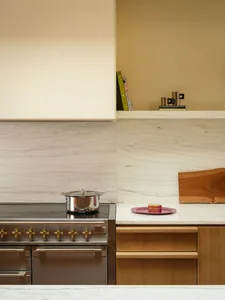 Modern kitchen corner featuring a stainless steel range with brass knobs, white marble backsplash and countertop, light oak cabinetry, and a wooden cutting board with colorful cookbooks on an upper shelf.
