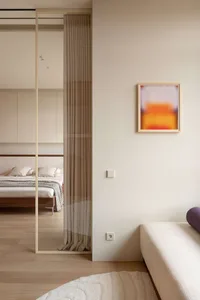 Modern minimalist bedroom viewed through a walnut-framed sliding glass partition with pleated curtain, featuring a low wooden bed with white bedding and abstract orange artwork on the adjacent wall.