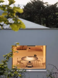 View from outside into a modern minimalist dining room with a round black table, white chairs, and warm lighting, framed by a concrete wall and green foliage.