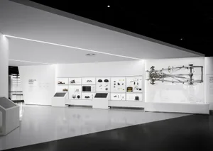 Modern minimalist exhibition hall with white walls displaying automotive parts, technical diagrams, and a large suspended car chassis on the right side.
