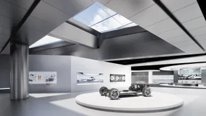 Modern automotive exhibition hall featuring a white circular platform displaying a car chassis, surrounded by technical displays and large skylights in a concrete and steel interior.