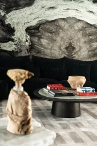 Modern living room featuring a large abstract green and grey marble wall, black velvet sectional sofa with multiple cushions, round dark coffee table with books and stone bowl, and foreground stone sculpture.