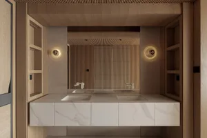 Modern minimalist bathroom with double white marble sinks set in a floating vanity, flanked by tall light oak shelving units and circular wall sconces, with a large mirror reflecting a wood slat accent wall.