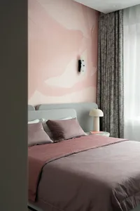 Modern bedroom with a large abstract blush pink mural wall, grey velvet headboard, dusty rose patterned bedding, and a sculptural white wall lamp.