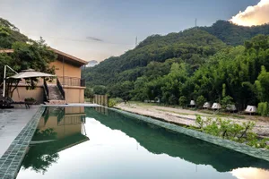 Long rectangular infinity pool with reflective green water, set beside a modern concrete villa with a wooden deck and lounge chairs, overlooking a lush green mountain valley with glamping tents in the distance.