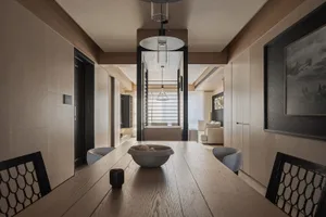Modern minimalist dining room featuring a long light oak table, black lattice-back chairs, wood panel walls, and a view through to a living area.