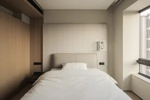 Modern minimalist bedroom with a beige upholstered bed, white linens, vertical wood panel accent wall, and large window with natural light.
