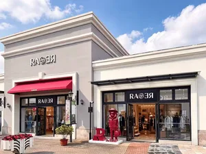 Exterior view of a RAOR fashion boutique with large glass windows, black frames, a bright red awning, and red bear sculptures on a sunny day.