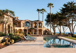 Luxurious Mediterranean-style stone villa with arched windows, terracotta roof tiles, and a long rectangular swimming pool surrounded by palm trees and ocean views.
