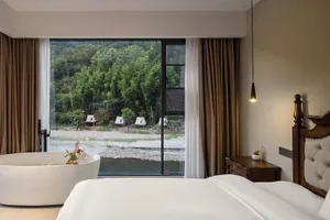 Modern hotel bedroom featuring a large white freestanding bathtub positioned directly in front of a floor-to-ceiling window overlooking a lush green forest and river, with a wooden four-poster bed and dark wood nightstand.