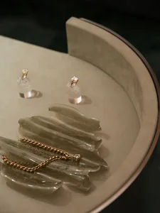 特寫半透明橄欖綠樹脂葉形盤，上面擺放金色麻花項鍊與愛心形鑽石墜飾，旁邊有兩枚鑽石 solitaire 戒指置於壓克力展示座上，整體放在弧形米色軟墊表面。