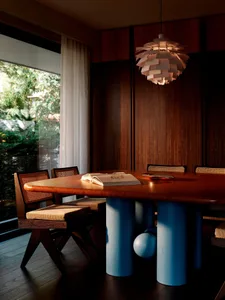 Modern dining room featuring a wooden table with bright blue cylindrical pedestal legs, surrounded by cane-back dining chairs and a large PH Artichoke pendant lamp, with walnut wood paneling and a large window overlooking greenery.