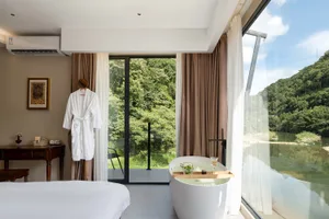 Modern hotel room with a white freestanding bathtub placed next to large glass windows offering a scenic view of a forested mountain and river.