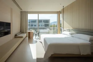 Modern minimalist hotel bedroom featuring light oak wood paneling, a large platform bed with white linens, built-in window seating, and a stunning mountain view through floor-to-ceiling glass doors.