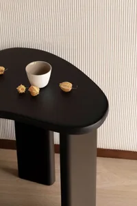 Close-up of a matte black console table with rounded edges, holding a white ceramic bowl and three dried physalis fruits against a vertically ribbed beige wall.