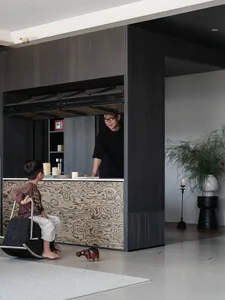 Modern dark-toned living room featuring a large burl wood patterned pass-through counter, a man standing behind the counter and a young boy sitting on a rocking chair in front, with a potted plant and candle on the right side.