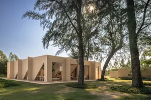 Exterior view of a modern beige villa featuring angular sandstone walls and large openings, surrounded by lush tropical trees and manicured lawn under a clear blue sky.