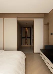 Modern minimalist bedroom interior featuring a large bed with white linens, floor-to-ceiling oak veneer wardrobes, herringbone wood flooring, and a view into a long hallway with black-framed doors.
