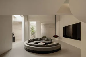 Modern minimalist living room featuring a large grey curved modular sofa on a circular platform, limestone walls, high ceilings with skylights, and a black pendant fireplace.