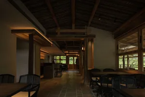 Interior view of a Japanese countryside villa dining room featuring dark wooden exposed beams, textured plaster walls, stone tile flooring, and black rattan dining chairs arranged around wooden tables.