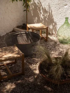 Sunlit Mediterranean courtyard featuring two wooden woven-rattan stools, a large galvanized metal tub, a terracotta planter with ornamental grass, a green glass demijohn, and a young citrus tree against a textured white stucco wall.