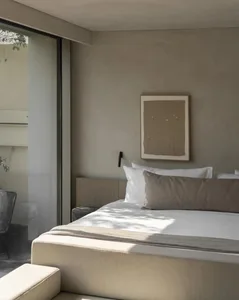 Minimalist bedroom interior featuring a low platform bed with white and beige linen bedding, a large textured beige artwork on a plaster wall, and soft natural light streaming through a floor-to-ceiling glass door.