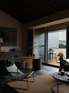 Modern living room featuring a dark wood ceiling, olive green sling chair, wooden console, and large glass doors opening to a balcony with mountain views.