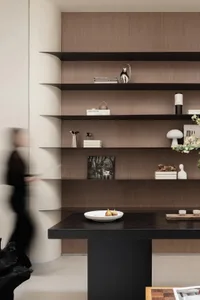 Modern living room with dark walnut floating shelves displaying books, sculptures, and decorative objects against a light neutral wall, with a black console table in the foreground.