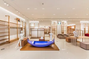 Modern fashion boutique interior featuring a glossy blue Venetian gondola display on a wooden platform, surrounded by minimalist oak shelving and white mannequins under bright track lighting.