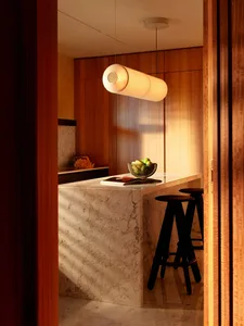 View into a modern kitchen featuring a white marble island with dark wood bar stools, warm walnut cabinetry, and a long cylindrical pendant light.