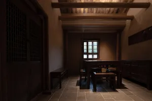 Interior view of a traditional Chinese dining room with dark wooden table, stools, side cabinet, exposed wooden ceiling beams, and a lattice door.