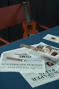 Blue table surface with scattered newspapers, washi tape, a wooden pencil, a brown leather chair, and a wooden ukulele in the background.