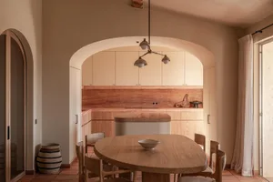 Warm minimalist dining room featuring a large oval oak dining table, wooden chairs, terracotta floor tiles, and an arched opening revealing a natural wood kitchen with a terracotta backsplash.