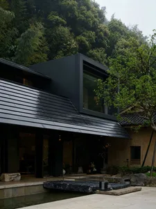 Modern black steel Japanese tea house with large glass windows, dark tiled roof, and stone water feature set against a dense green forest.
