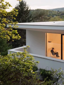 Modern minimalist concrete house with large opening to interior, man reading on wire chair, surrounded by lush green trees and forest view at dusk.
