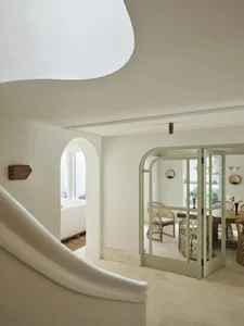View from a curved staircase into a bright dining room with sage green arched French doors, rattan dining chairs, and white plaster walls.