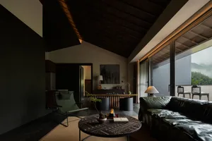 Modern living room featuring a dark wood ceiling and walls, a dark green leather sofa, a matching green lounge chair, an intricately carved round coffee table, and large windows overlooking misty mountains.