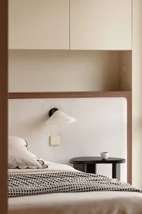 Modern minimalist bedroom with a cream-colored vertically ribbed fabric headboard, black cone-shaped wall sconce, dark oval bedside table, and neutral bedding with black-and-white checkered throw.