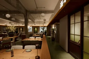 Interior view of a modern Japanese restaurant featuring long light oak dining tables, upholstered chairs, exposed concrete beams, and hanging spherical pendant lights.