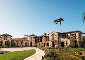 Luxurious Mediterranean-style villa with light-colored stone masonry, arched windows, terracotta roof, and a landscaped courtyard with palm trees under a clear blue sky.