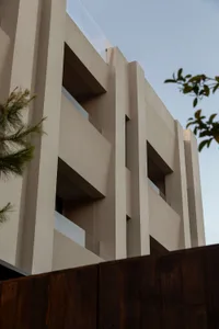 Low-angle view of a modern multi-story concrete building featuring protruding structural elements, recessed balconies with glass railings, and a dark wooden fence in the foreground with tree branches on the sides.